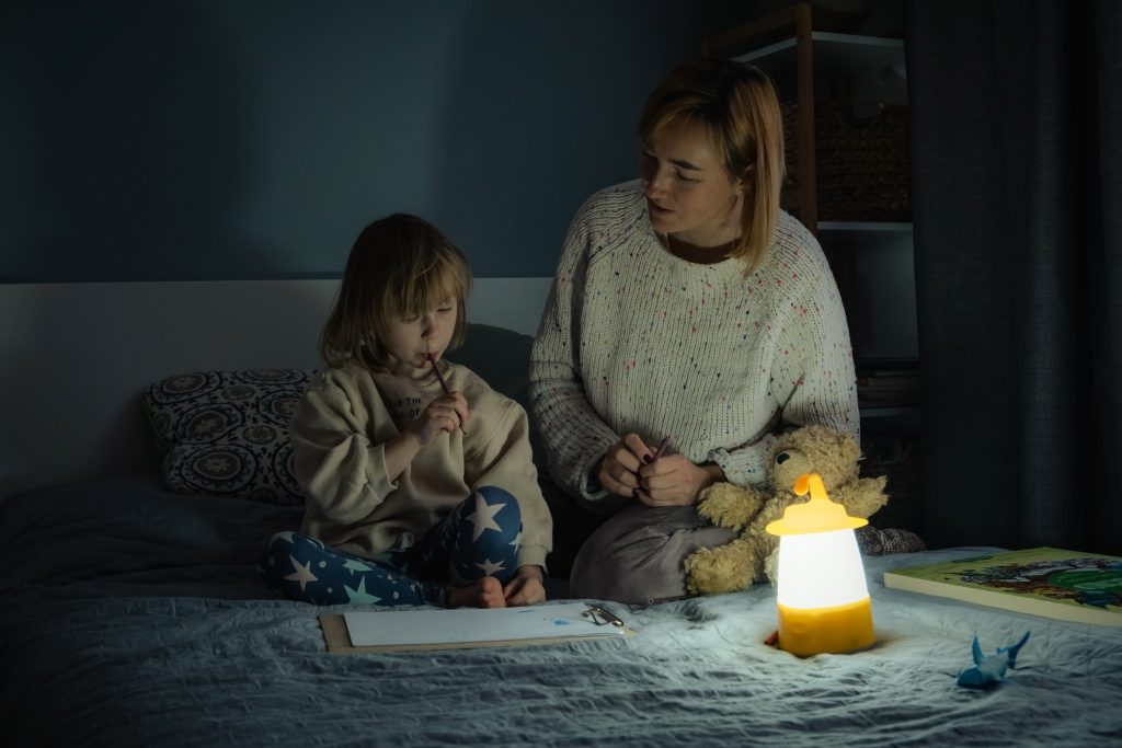 Mother And Little Daughter Studying And Drawing In A Complete Darkness During Electricity Outage. Little Girl Uses Camping Lantern To Do Her Homework During Blackout. Energy Crisis Concept